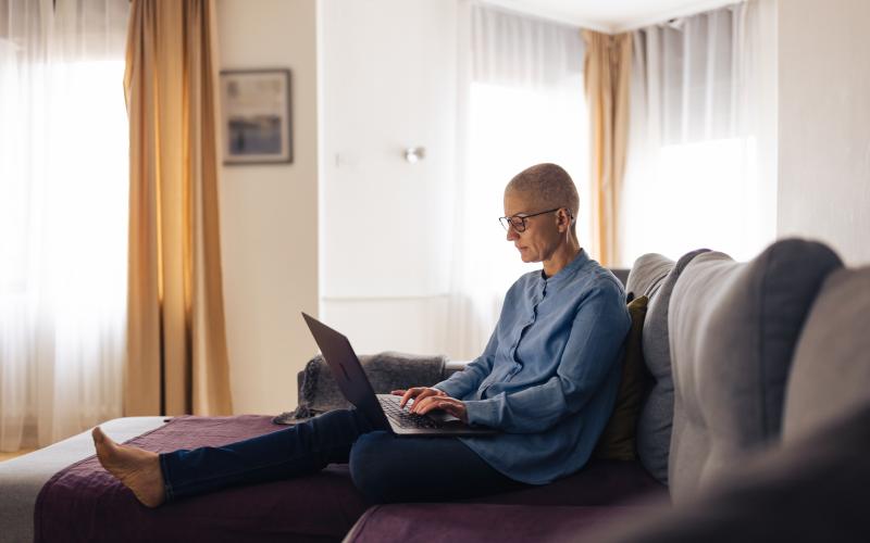 Een vrouw zonder haar zit met haar laptop op schoot in haar woonkamer op de bank.