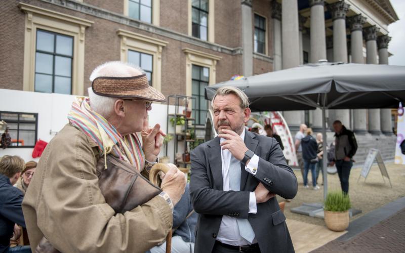 Nationale ombudsman Reinier van Zutphen in gesprek met een oudere meneer. Ze staan op een druk plein met op de achtergrond andere mensen die in gesprek zijn.