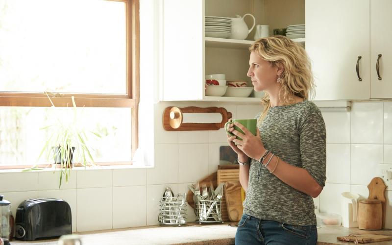 Een vrouw met blond krullend haar staat in haar keuken en kijkt uit het raam. In haar hand heeft ze een mok.