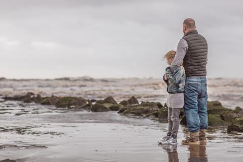 Man staat samen met kind op het strand. Ze kijken naar de zee