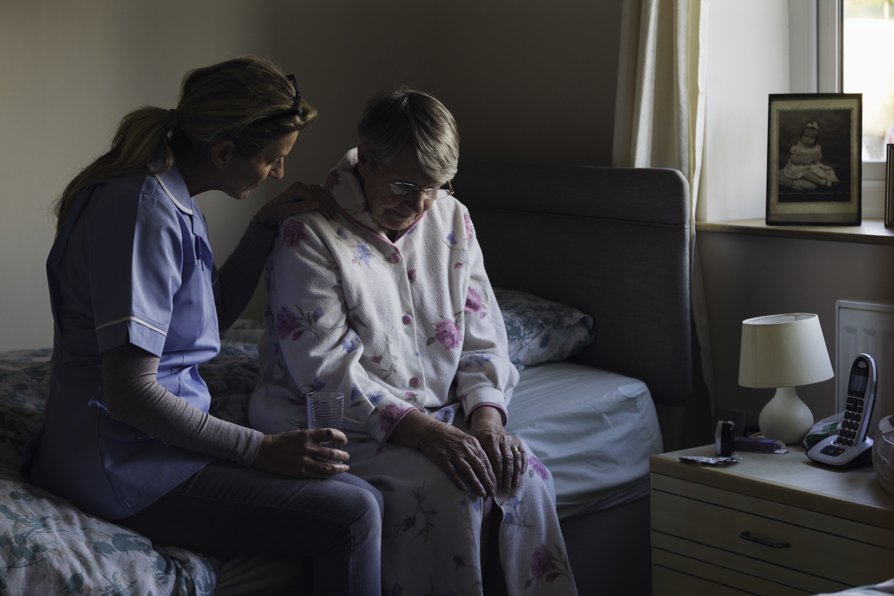 Foto van een zieke vrouw die op bed zit samen met een verpleegster. De verpleegster slaat een arm om de vrouw heen.