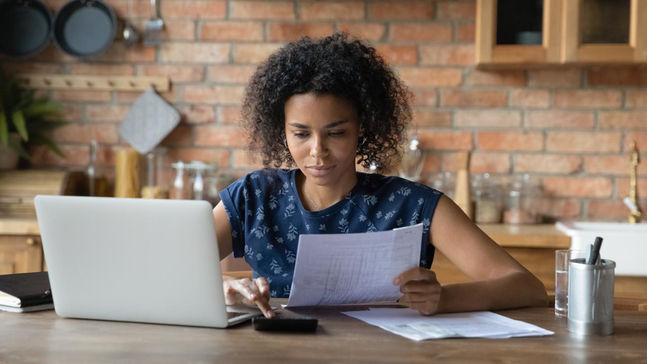 Vrouw zit aan tafel met papier en laptop voor zich en regelt financiën