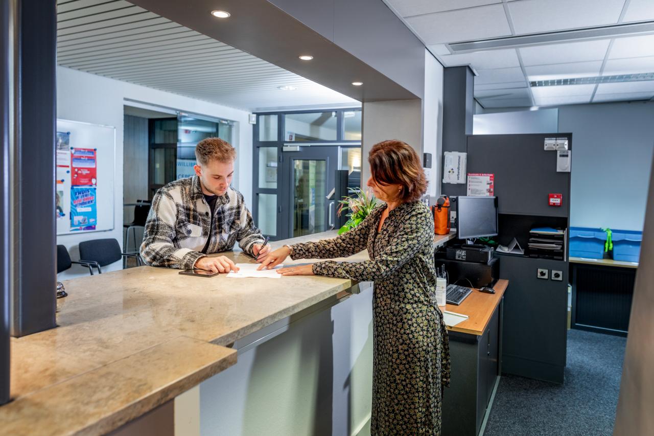 Jongere man staat bij de gemeente aan de balie. De vrouw achter de balie laat hem een formulier zien.