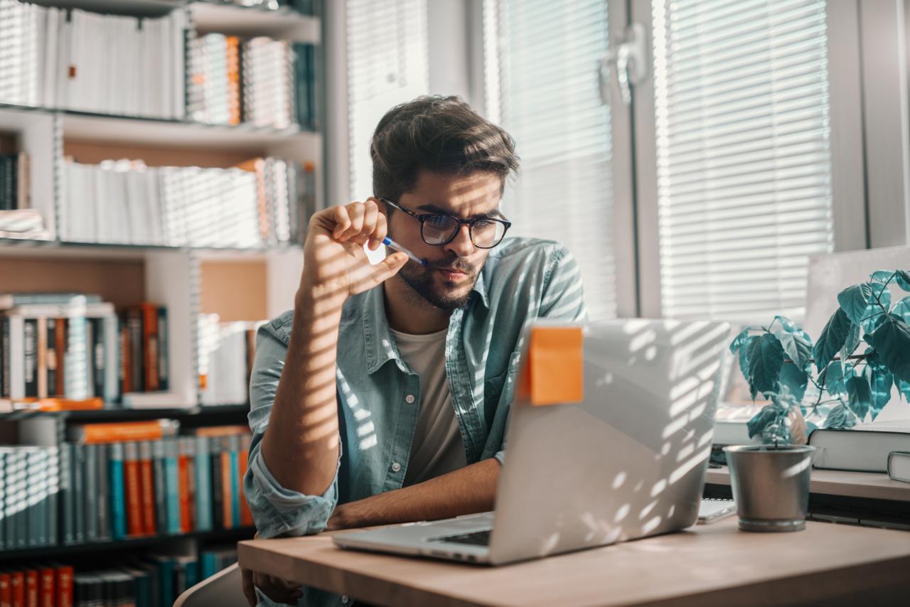 Een jongvolwassen man zit in een bibliotheek en kijkt naar zijn laptopscherm