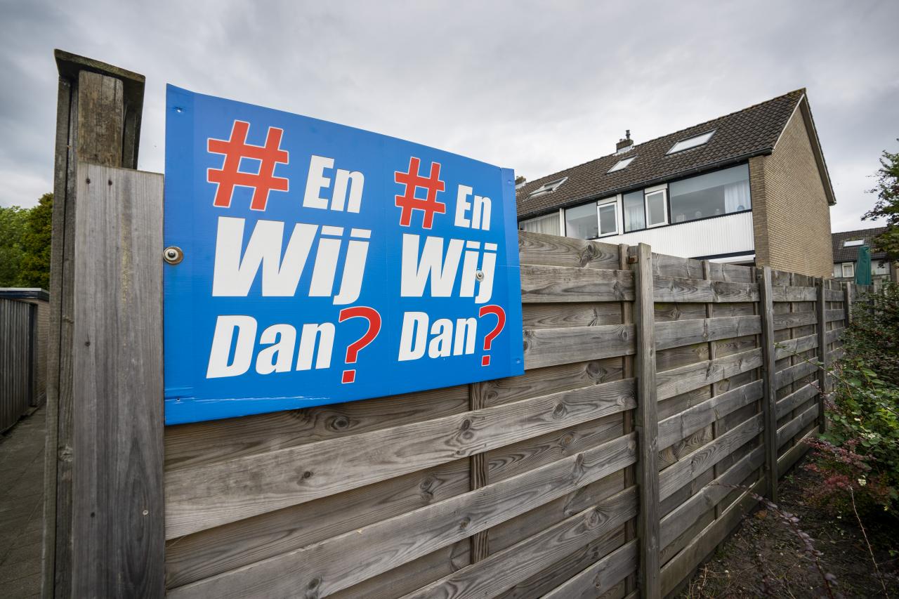 Bord hangt aan tuinhek met daarop de tekst En Wij Dan?