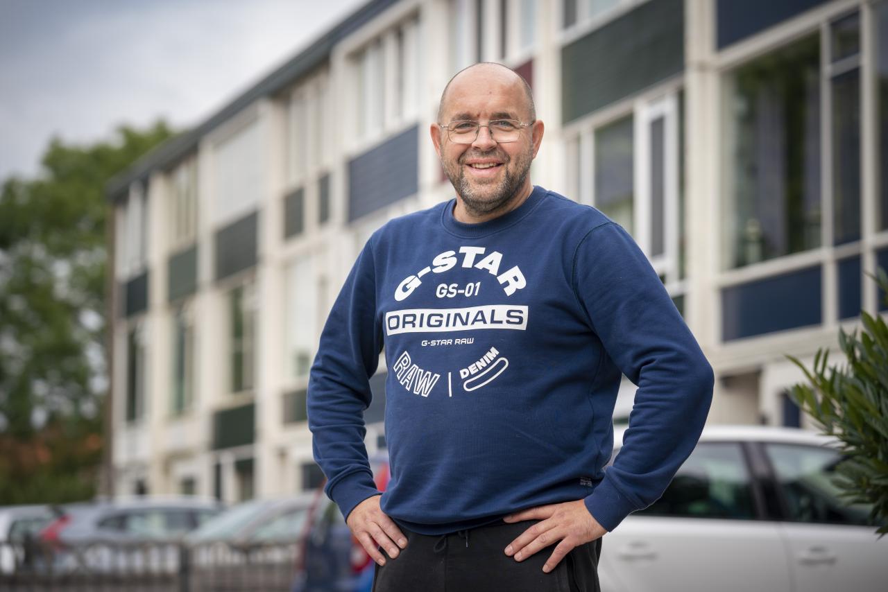 Man met bril en blauwe trui staat lachend voor een blok huizen