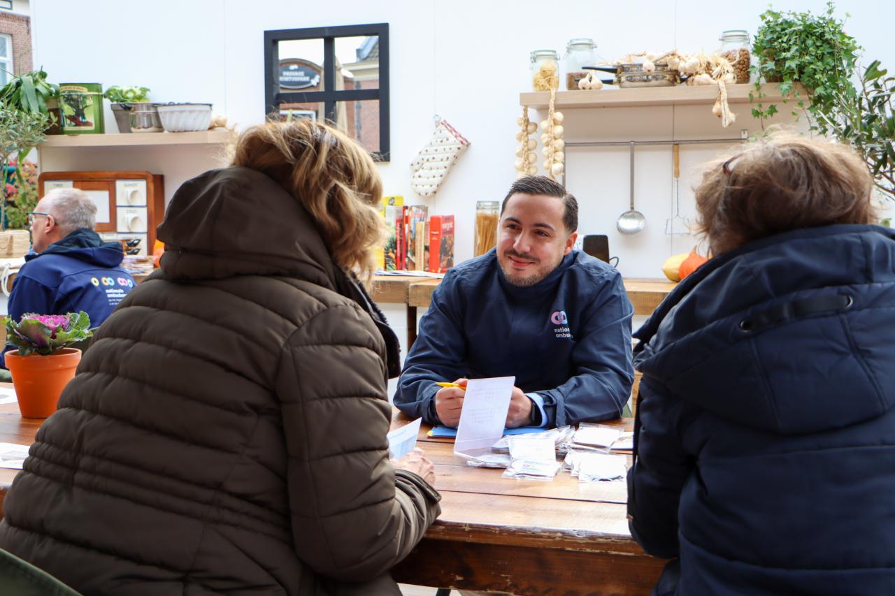 Een jongvolwassen man met stoppelbaardje en snor en zwart haar, werkzaam bij de Nationale ombudsman is in gesprek met twee vrouwen op straat.