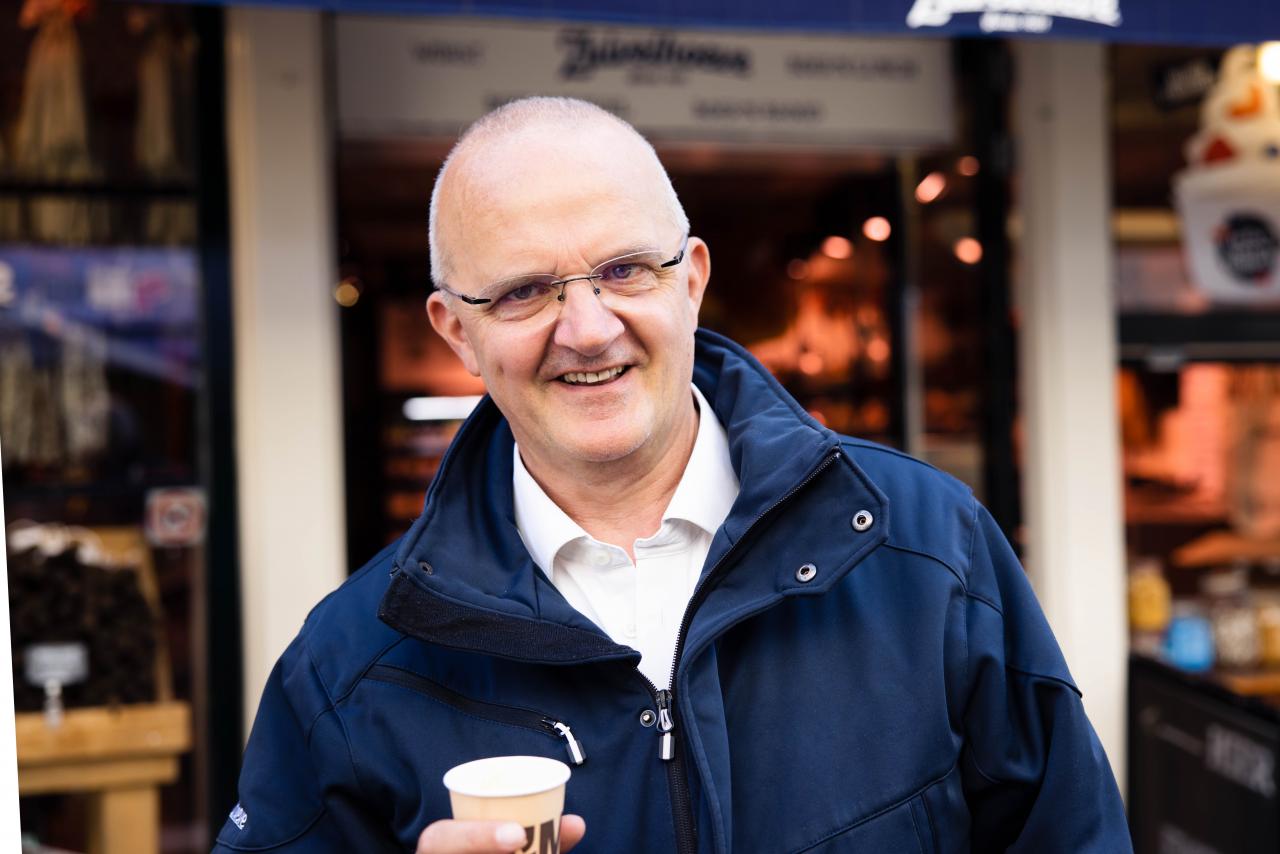 Anton, een kale man met bril van 58 kijkt in de camera en glimlacht. Hij houdt een beker koffie vast.