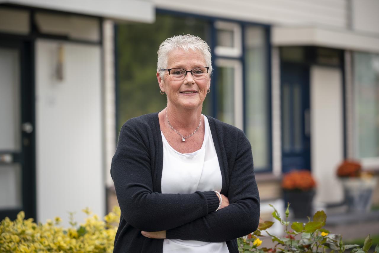 Vrouw met kort grijs haar, zwarte bril en zwart vest en wit t-shirt staat voor haar huis. Ze heeft haar armen over elkaar gekruist.