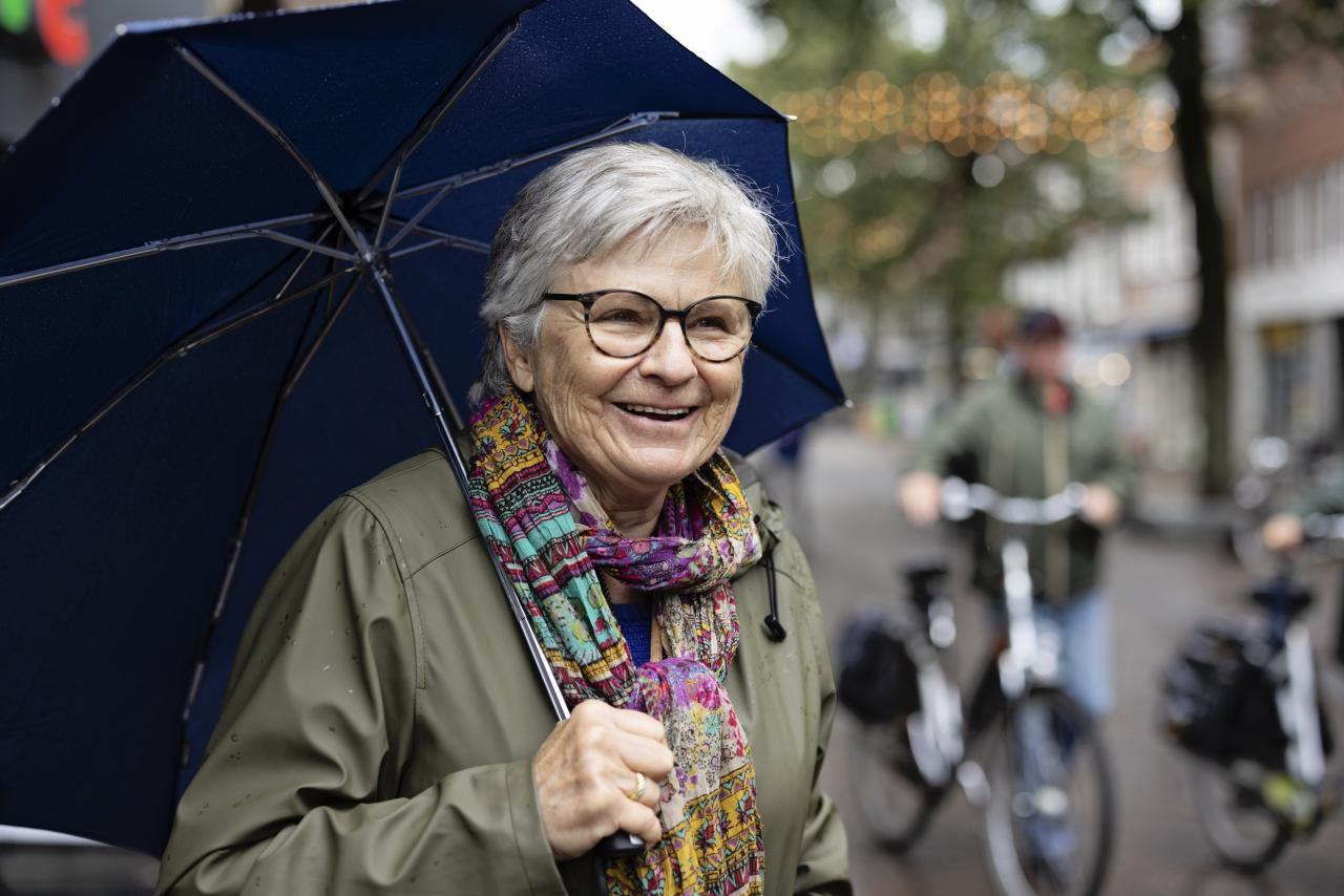 Marga, een oudere vrouw met grijs haar, bril en een gekleurde sjaal om. Ze heeft een paraplu vast