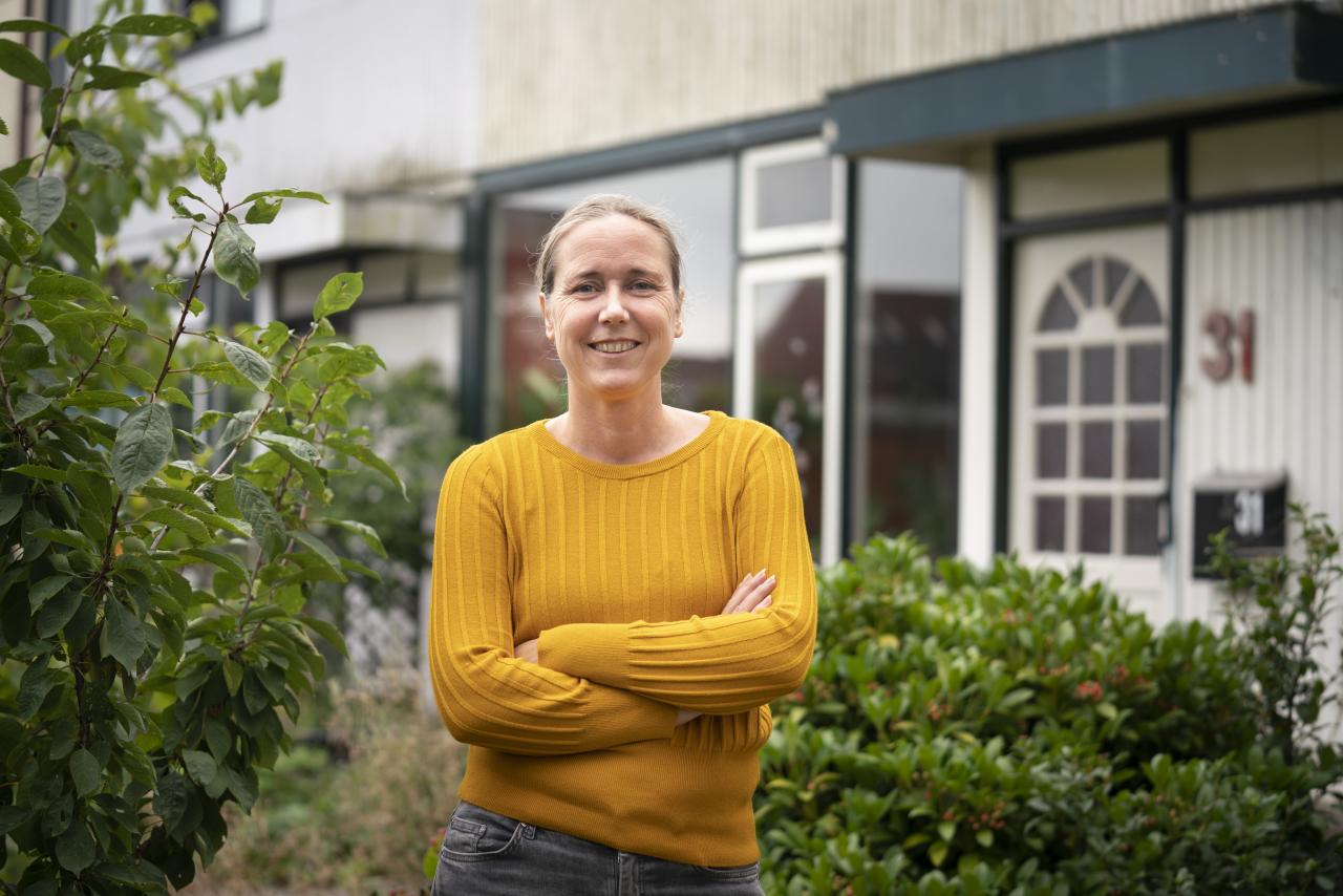 Vrouw met gele trui staat met armen over elkaar voor een woning