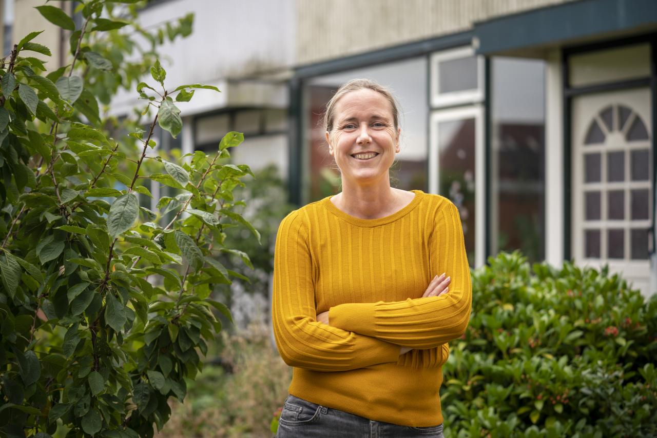 Vrouw kijkt lachend in de camera. Ze staat voor haar huis, heeft een gele trui aan en houdt haar armen over elkaar gekruist