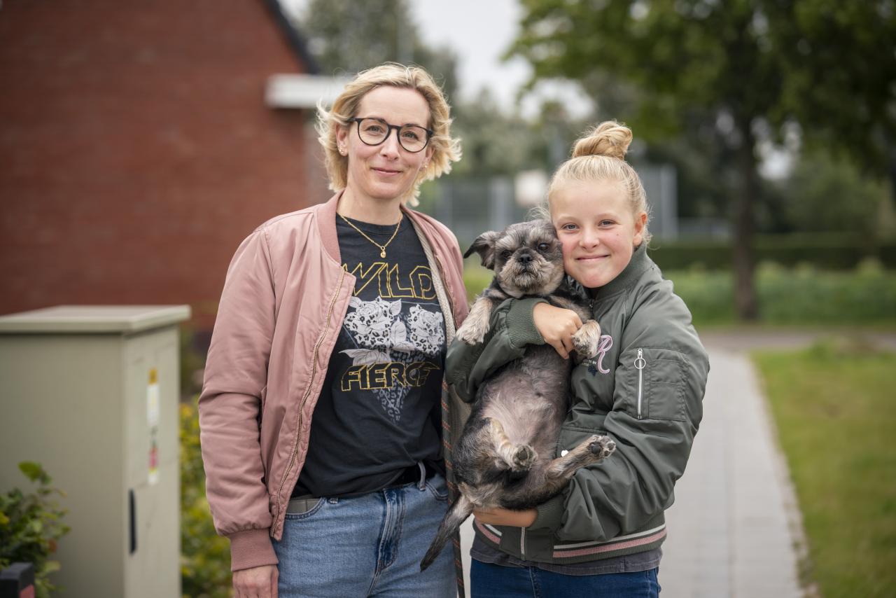 Vrouw met blond haar, zwarte bril en roze jas staat samen met haar dochten (blond haar in knot en draagt groene jas) op straat. Ze houden een hond in de hand.