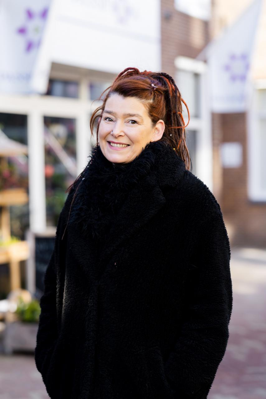 Miranda, een vrouw van middelbare leeftijd met rood haar en dreadlocks. Ze draagt een zwarte jas.