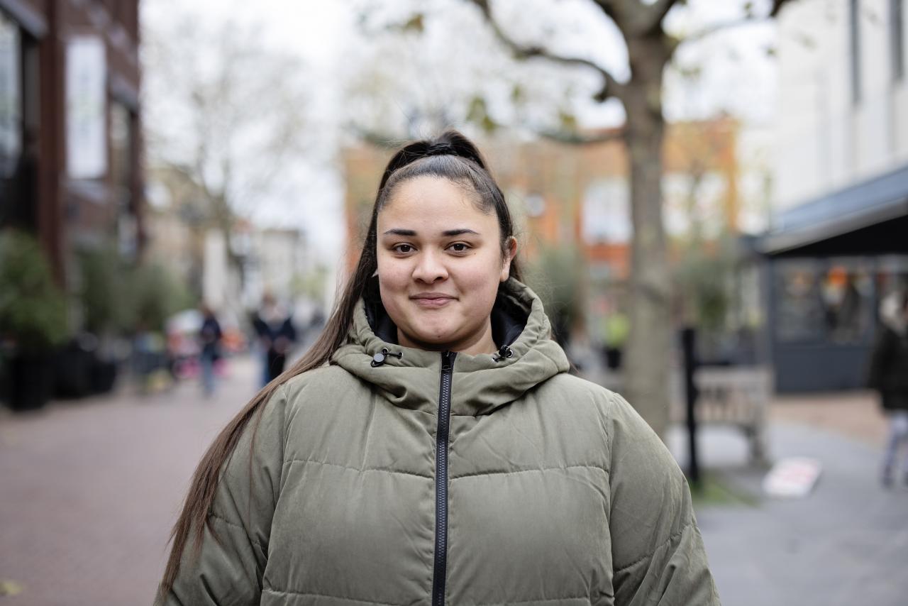 Jonge vrouw (student) met lang donkerbruin haar in een staart en een groene winterjas kijkt in de camera.