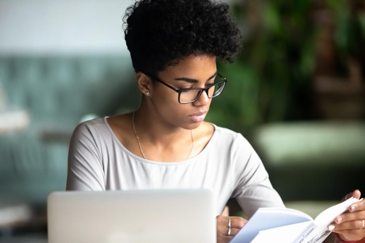 Een vrouw met zwart kroes haar zit achter haar laptop heeft een boek vast