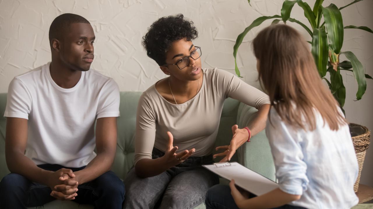 Een jonge vrouw en haar vriend zitten op de bank en leggen iets uit aan een andere vrouw, die met een kladblok luistert. 