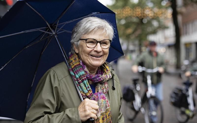 Marga, een oudere vrouw met grijs haar, bril en een gekleurde sjaal om. Ze heeft een paraplu vast