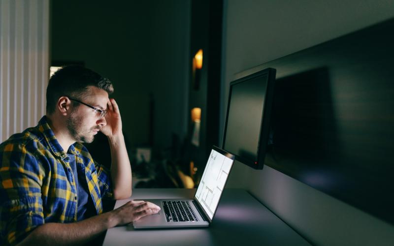 Man met bril zit achter computer. Zijn hand ondersteund zijn hoofd. De omgeving is donker.