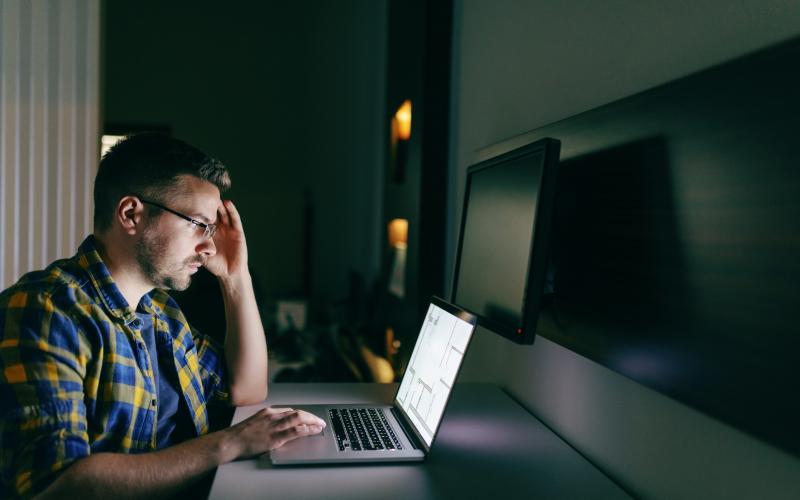 Man met bril zit achter computer. Zijn hand ondersteund zijn hoofd. De omgeving is donker. Man met bril zit achter computer. Zijn hand ondersteund zijn hoofd. De omgeving is donker.