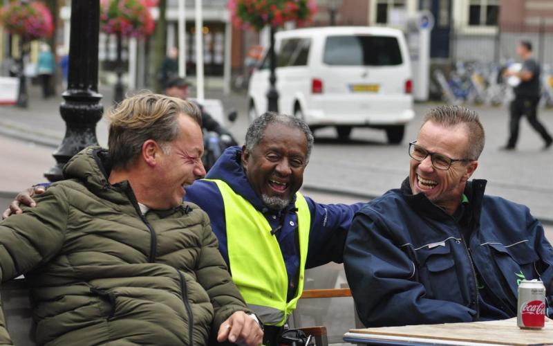 Drie mannen zitten op een terras. Ze lachen naar elkaar.