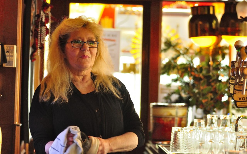 Vrouw van middelbare leeftijd staat achter de bar in een kroeg en maakt een glas schoon.