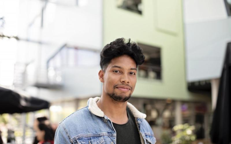 Een jongvolwassen man met baardje en spijkerjack aan. Hij staat in een winkelcentrum.