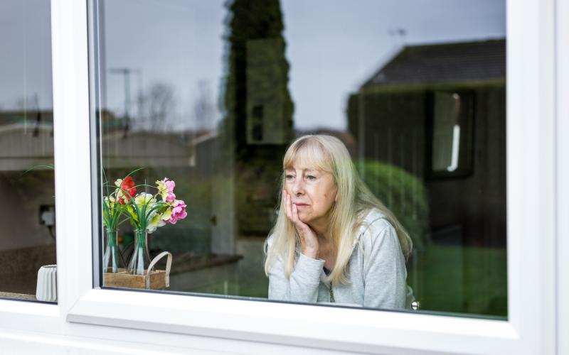 Oudere vrouw zit achter het raam en kijkt naar buiten. Ze kijkt zorgelijk en steunt haar hoofd op haar hand.