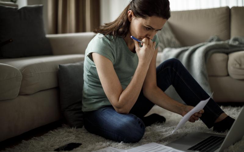 Vrouw zit op de grond en heeft papieren in haar hand. Voor haar staat ook een laptop.