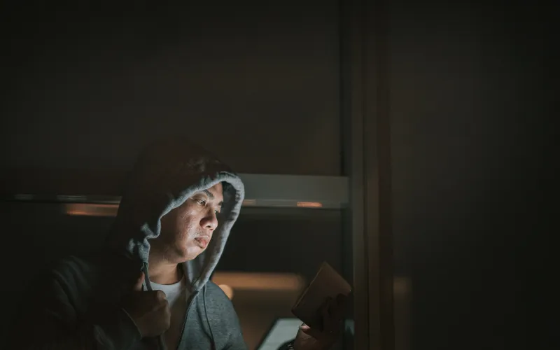 Een man zit in een donkere kamer, heeft zijn capuchon van zijn hoodie op en kijkt naar een tablet. Zijn gezicht is zichtbaar door het licht van de tablet.