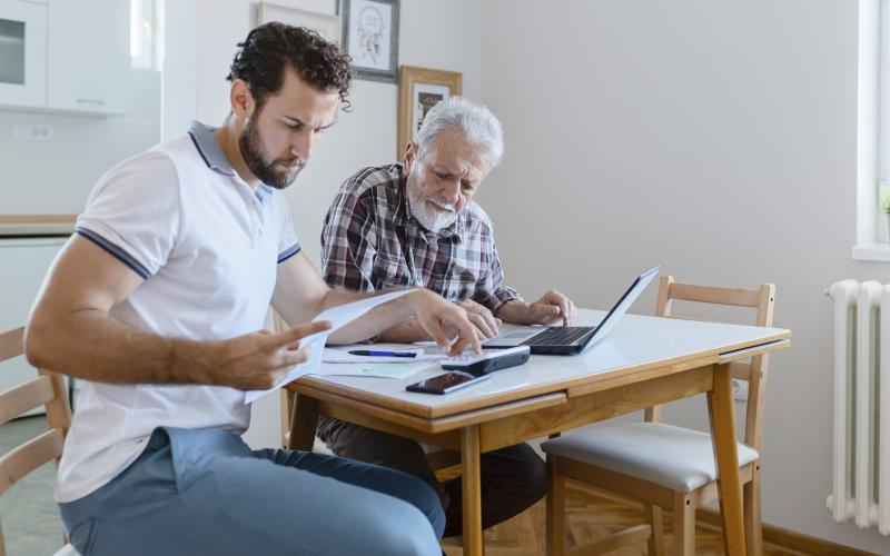 Een jongvolwassen man en senior man kijken samen naar de financiën. Ze hebben papierwerk, een rekenmachine en een laptop voor zich op tafel.