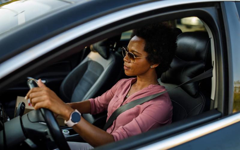Een vrouw zit in een auto achter het stuur. Ze draagt een zonnebril en kijkt voor zich uit naar de weg.