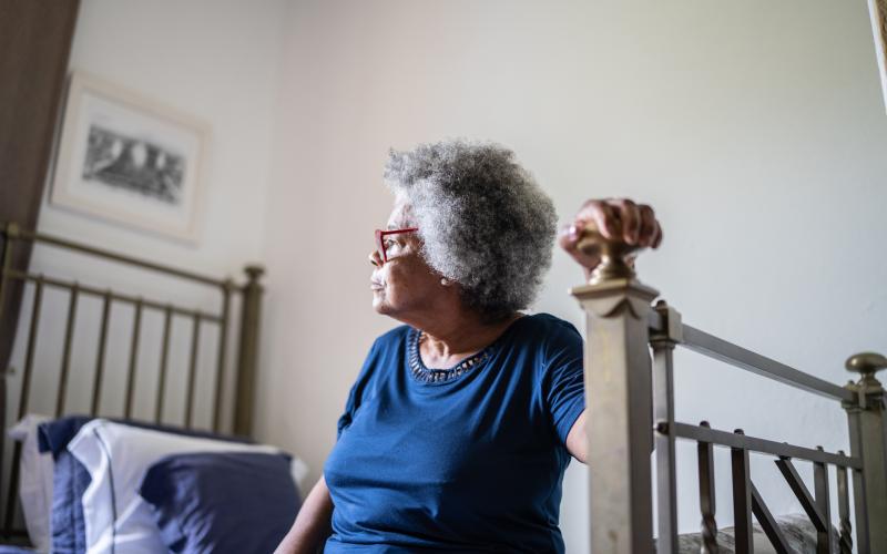 Senior vrouw met bril en blauw t-shirt zit op haar bed. Ze houdt met één hand het bedframe vast om op te gaan staan.