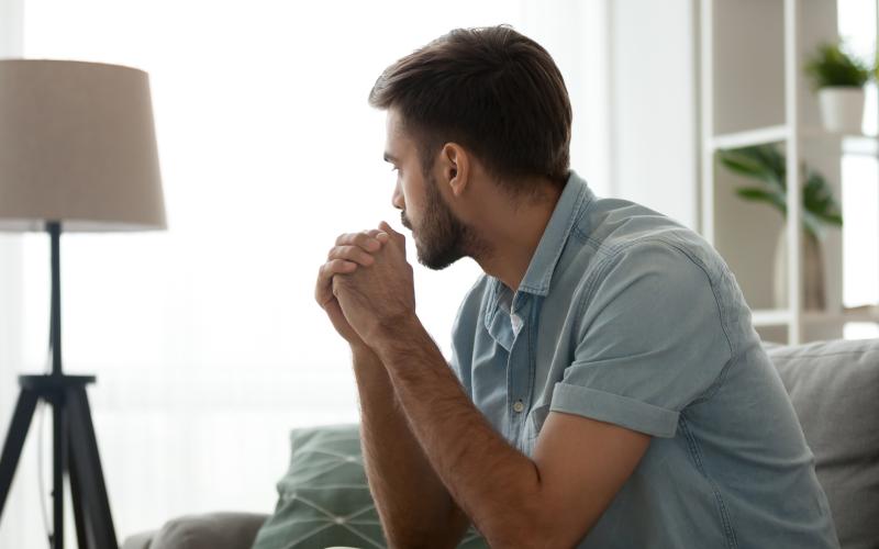 Een man van middelbare leeftijd met donker haar en een blauwe blouse zit op de bank. Hij kijkt nadenkend voor zich uit.