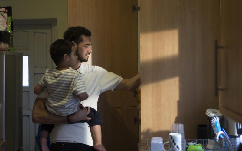 Man draagt zoontje op zijn arm en pakt tegelijkertijd iets uit een keukenkastje.