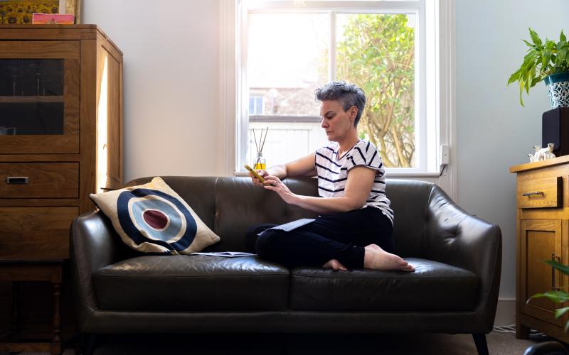 Vrouw zit thuis op haar bank voor het raam. Ze heeft haar telefoon vast en kijkt bezorgd. Vrouw zit thuis op haar bank voor het raam. Ze heeft haar telefoon vast en kijkt bezorgd.