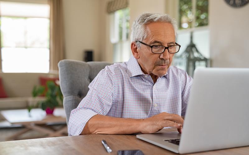 Oudere man (grijs haar, bril en geruit overhemd) werkt op een laptop. Hij zit in een woonkamer. Oudere man (grijs haar, bril en geruit overhemd) werkt op een laptop. Hij zit in een woonkamer.