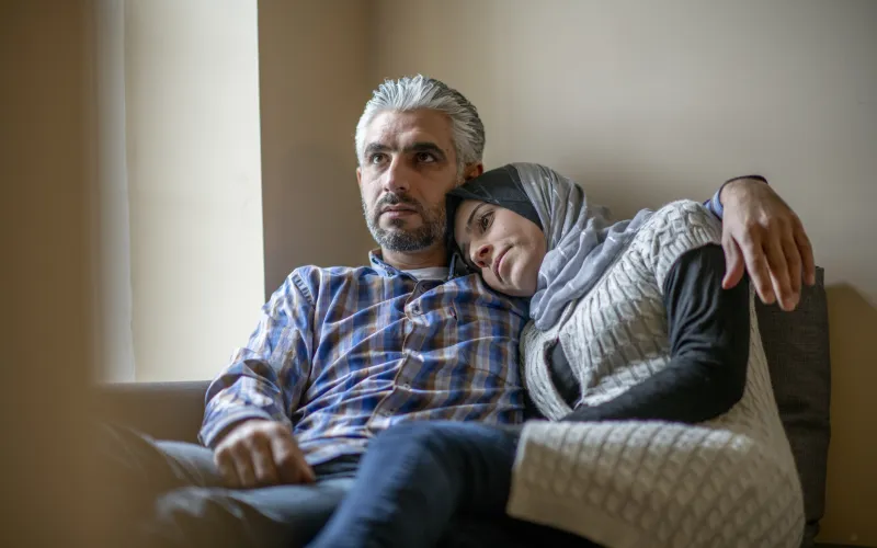 Een man en vrouw van islamitische afkomst zitten samen op de bank. Ze kijken bezorgd voor zich uit.