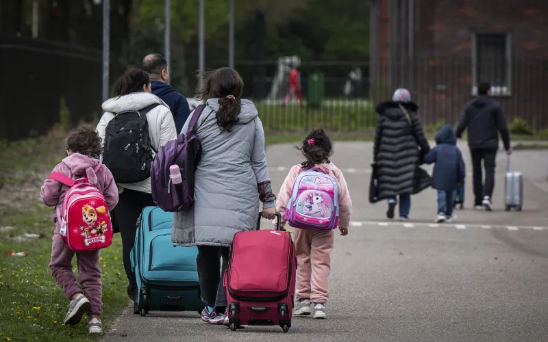 Groep mannen, vrouwen en kinderen lopen bij opvanglocatie. Ze dragen warme jassen, tassen en een rugzak. 