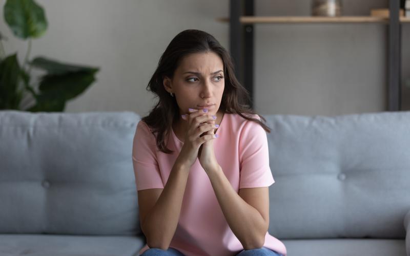 Vrouw met lang donker haar zit op de bank. Ze kijkt zorgelijk voor zich uit.