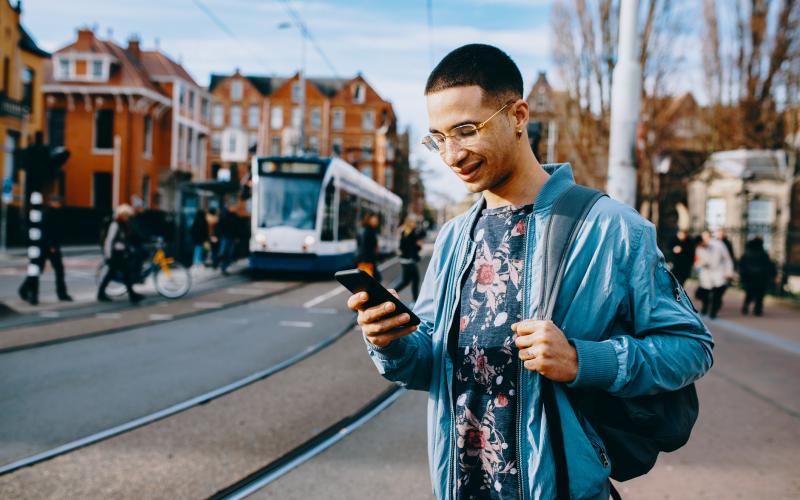 Een jongvolwassen man staat op straat met zijn telefoon in zijn hand. Op de achtergrond zie je mensen lopen en een tram.