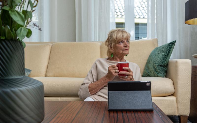 Oudere vrouw zit thuis op de grond voor haar bank. Ze kijkt opzij en houdt een kopje koffie vast.