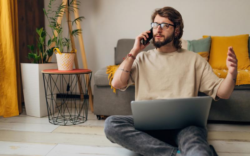 Jongvolwassen man zit op de grond voor zijn bank. Hij is aan het bellen en heeft zijn laptop op schoot. Jongvolwassen man zit op de grond voor zijn bank. Hij is aan het bellen en heeft zijn laptop op schoot.