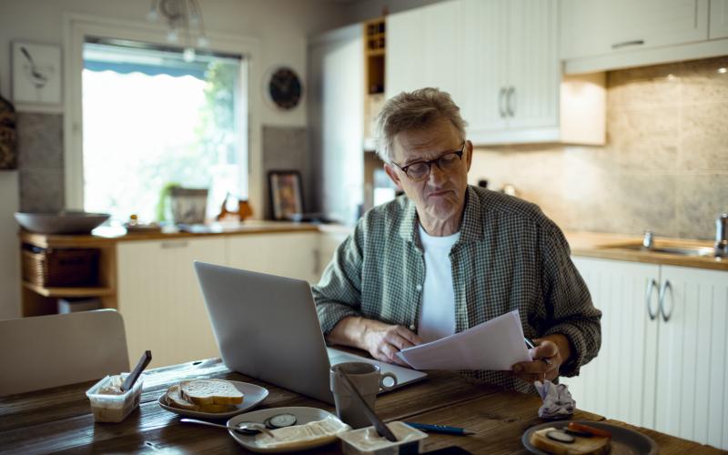 Senior man zit aan keukentafel met brief rekening in hand en laptop voor zich