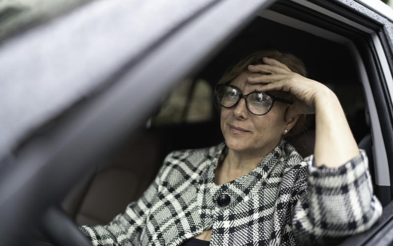 Een vrouw zit in een auto achter het stuur. Ze draagt een bril en geruit jasje.