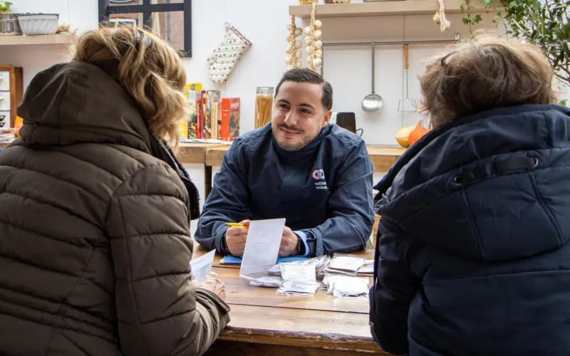 Een medewerker van de Nationale ombudsman in gesprek met twee mensen. Ze zitten aan een keukentafel.