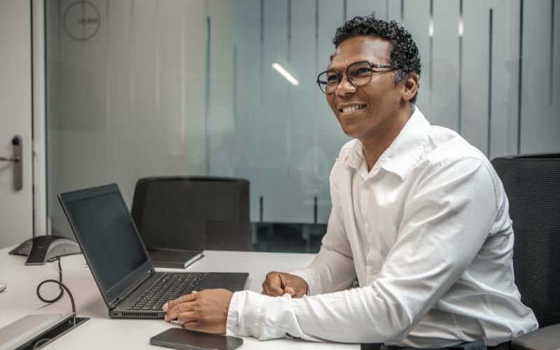 Man achter laptop aan het werk, hij kijkt vriendelijk op