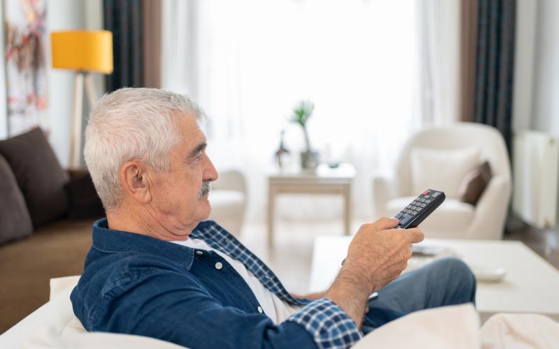 Een senior man zit op zijn bank voor de televisie. Hij heeft een afstandsbediening in zijn hand.