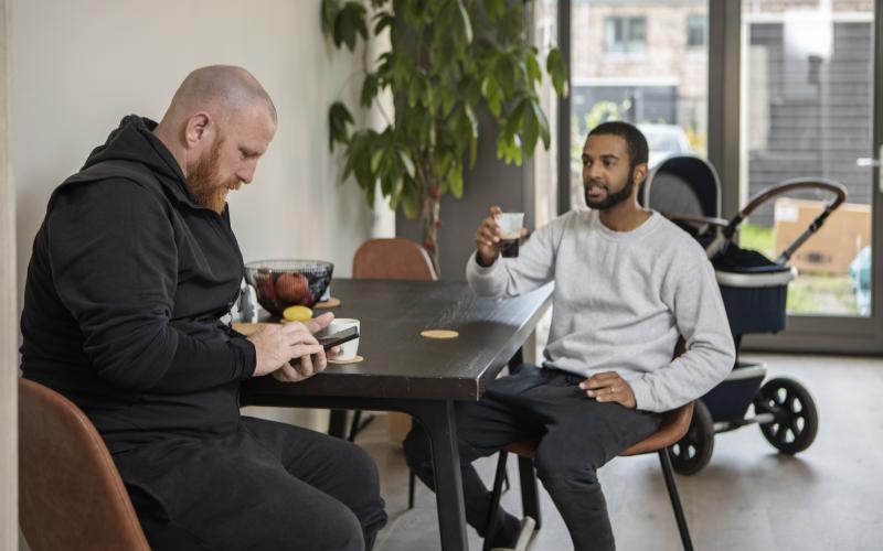 Twee mannen zitten aan een tafel. In de kamer staat een plant en kinderwagen