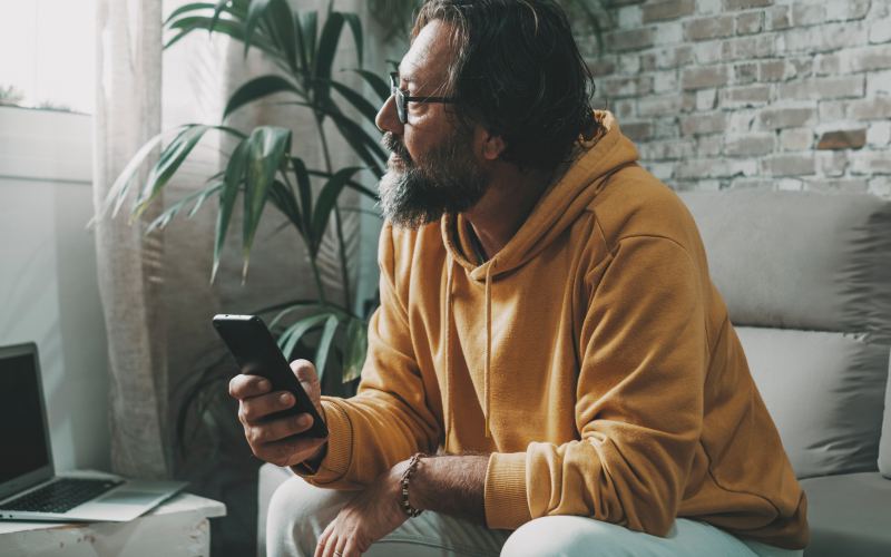 Man zit op de bank met telefoon in zijn hand. Hij kijkt naar buiten.